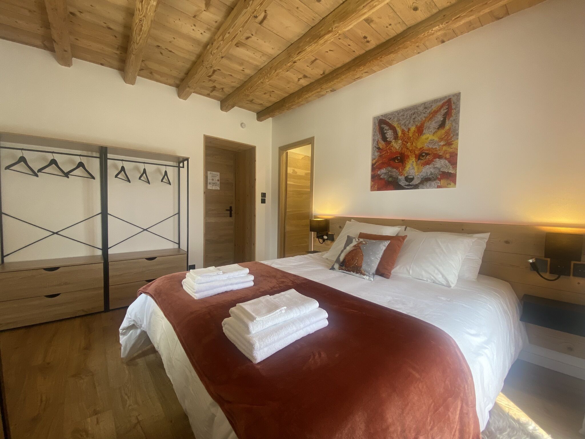 Une chambre d’hôtel avec un plafond en bois, un lit avec du linge de maison blanc et une couverture rouge, un tableau représentant un renard accroché au mur, et une armoire en bois avec des cintres.