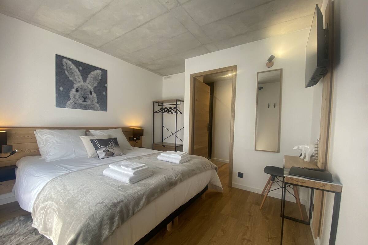 A modern hotel room with a large bed, a bedside table with a lamp, a wall-mounted TV, and a wooden floor.
