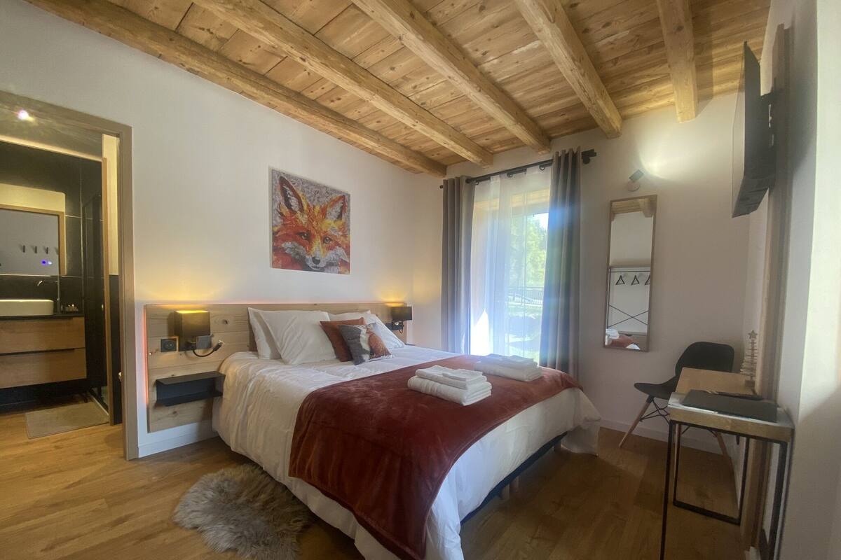 A bedroom with a wooden ceiling, a bed with a red blanket, a painting of a fox, a window with curtains, and a small table with a chair.