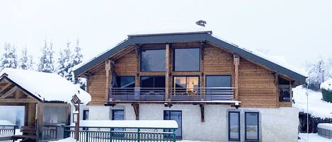 A wooden chalet with a balcony, large windows, and a snow-covered roof.