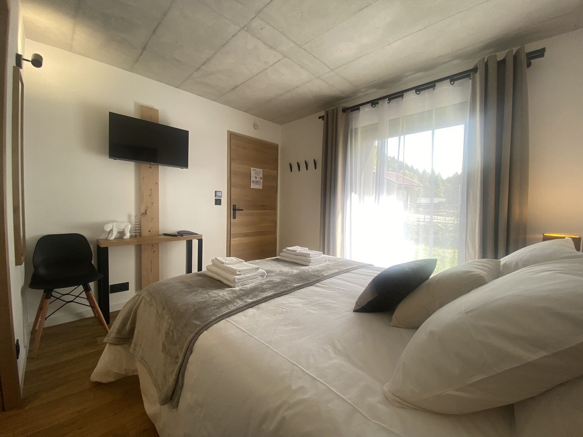 A hotel room with a bed, a desk, a chair, a TV mounted on the wall, and a window with curtains.