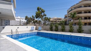 A rectangular swimming pool with a clear blue water surface, surrounded by a white tiled deck. There are two lounge chairs with striped cushions and a small palm tree in the background. - Maryam's 2-BR Apt Facing Larnaca Harbor W/ Pool (Cyprus - Larnaca)