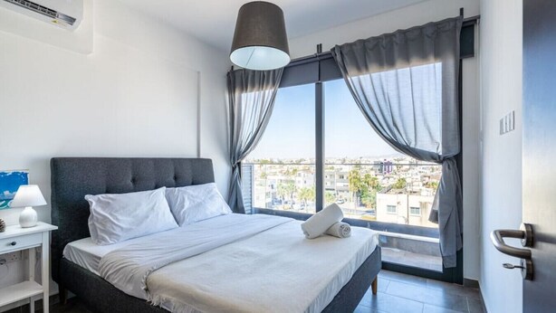 A modern hotel room with a large bed, a bedside table with a lamp, a window with curtains, and a city view. - Maryam's 2-BR Apt Facing Larnaca Harbor W/ Pool (Cyprus - Larnaca)