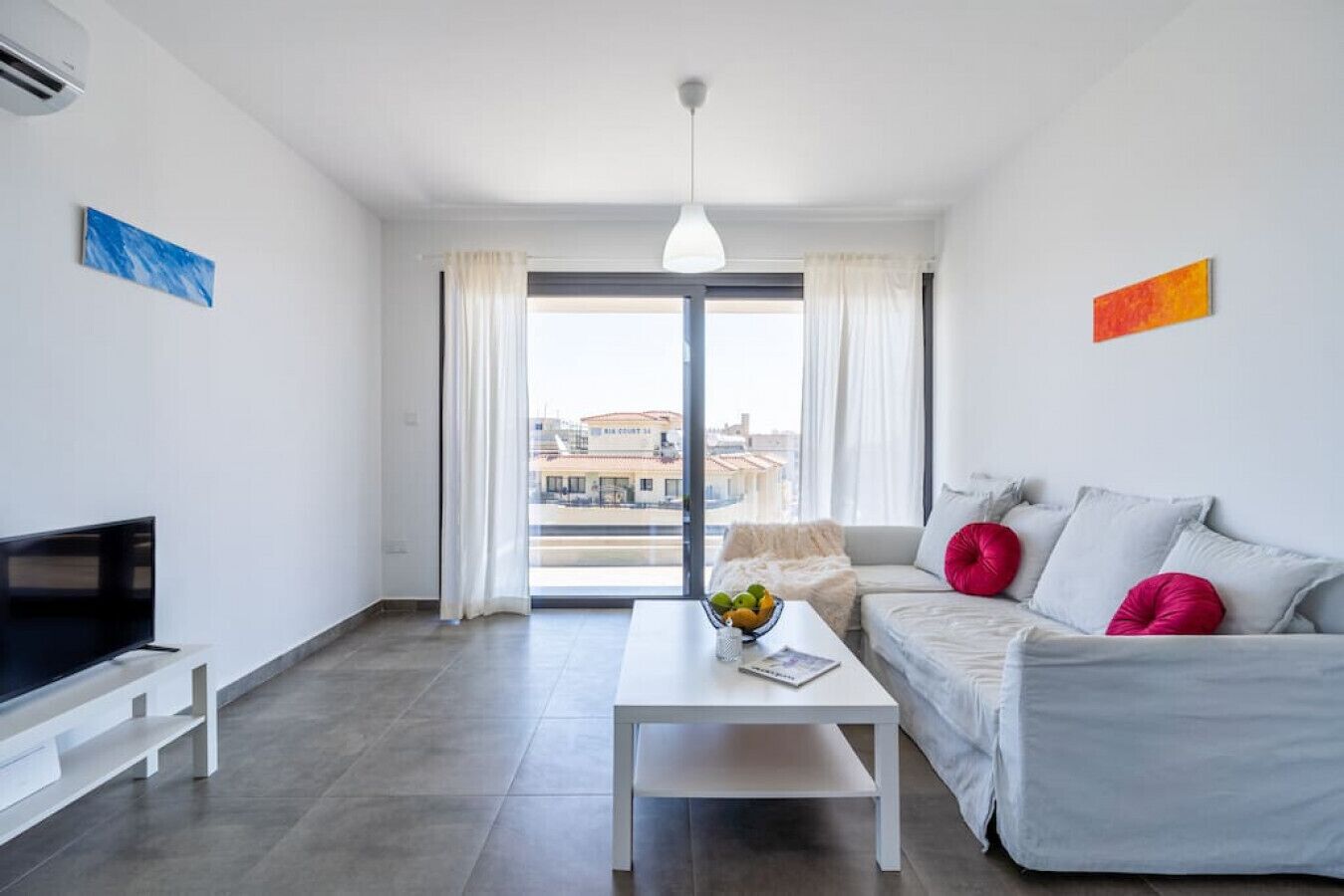 Una sala moderna con un televisor de pantalla plana, una mesa de centro blanca con un frutero, un sofá gris con cojines rojos y un ventanal con cortinas.
