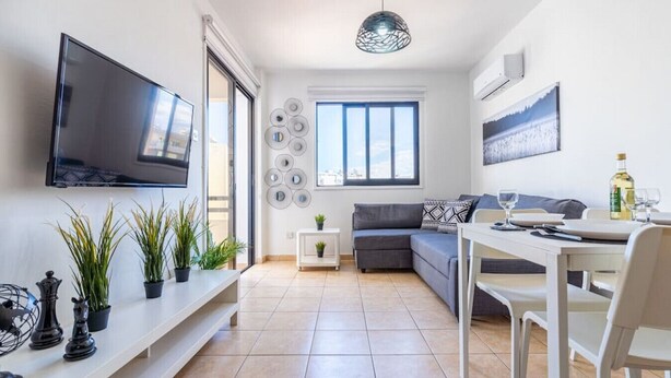 A modern living room with a flat-screen TV, a dining area with a table and chairs, a gray sofa, and a small side table with a plant.