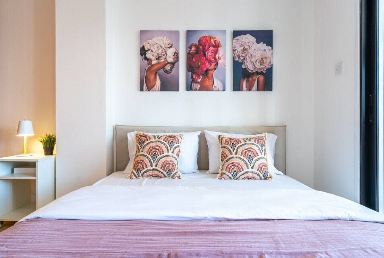 A neatly made bed with patterned pillows, a bedside table with a lamp, and three floral paintings on the wall.