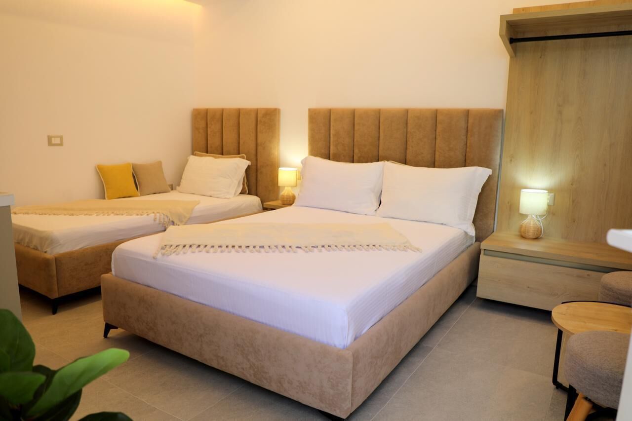 A hotel room with two beds, each with a headboard, a bedside table with a lamp, and a small round table with a chair.