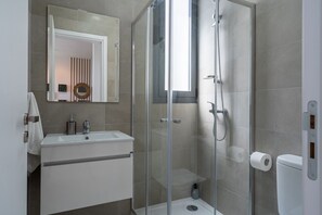 A modern bathroom with a glass-enclosed shower, a white sink with a mirror above, and a toilet with a paper towel holder. - Ivory 1-Bedroom Apartment in Aradipou (Cyprus - Larnaca)