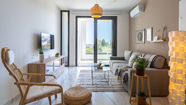 Ein modernes Wohnzimmer mit einer Couch, einem Korbstuhl, einem Couchtisch und einem Fernseher. Ein großes Fenster bietet Blick ins Freie.