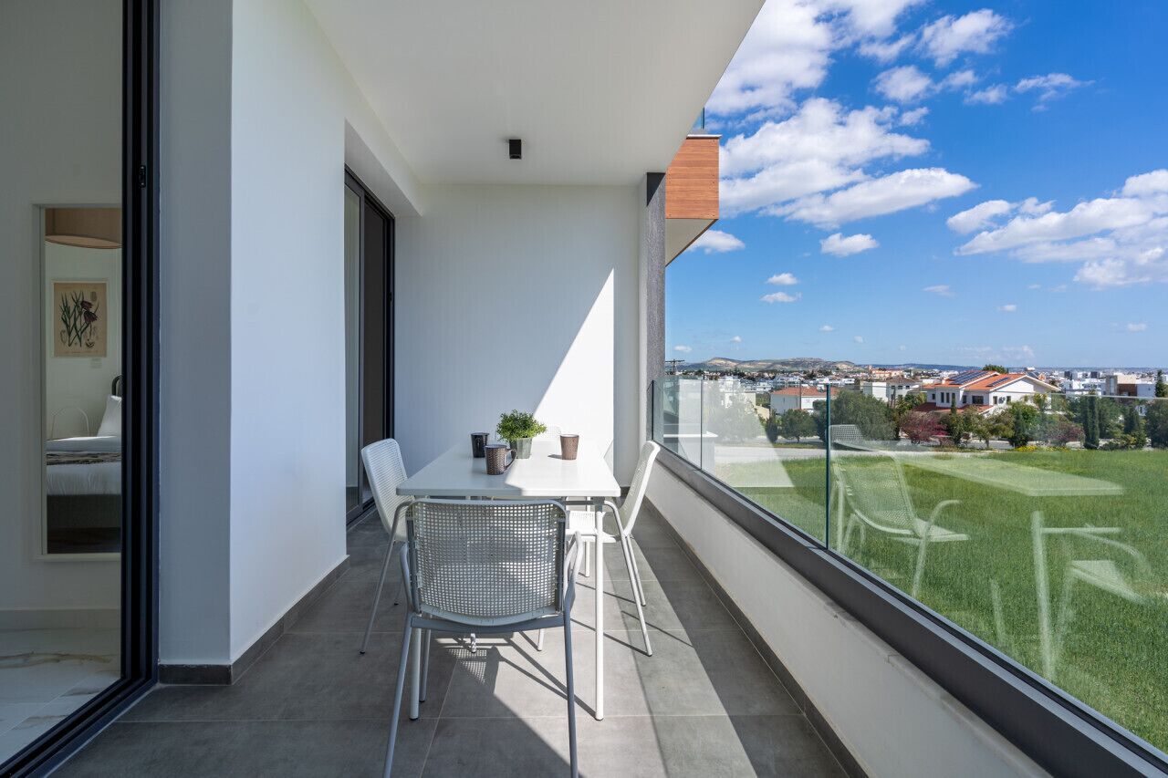A balcony with a table and chairs, a potted plant, and a view of a green landscape and buildings.
