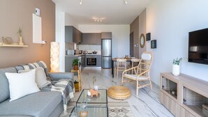 Ein modernes Wohnzimmer mit einem grauen Sofa, einem Glastisch und einem Korbstuhl. Im Hintergrund ist eine Küche mit grauen Schränken und einem Edelstahlkühlschrank zu sehen.