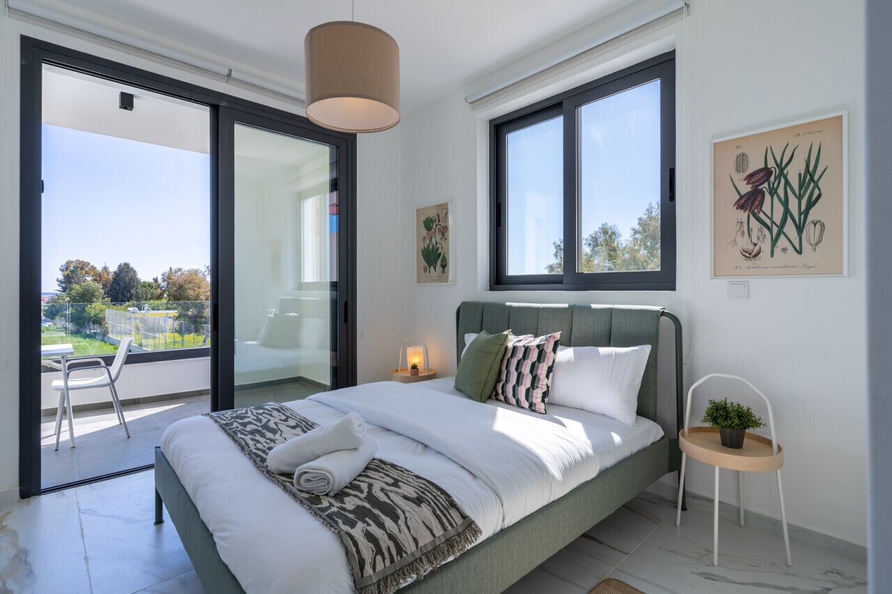 A bedroom with a bed, a bedside table, a chair, a window, and a balcony.