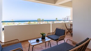 A balcony with wicker furniture, a glass table, and a view of the sea and buildings. - Bayview 2-BR Apartment in Larnaca (Cyprus - Larnaca)