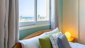 A hotel room with a bed, a nightstand with a lamp, and a window with a view of the beach and palm trees. - Bayview 2-BR Apartment in Larnaca (Cyprus - Larnaca)