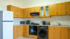 A compact kitchen with wooden cabinets, a refrigerator, a microwave, an oven, a washing machine, and a coffee maker. - Bayview 2-BR Apartment in Larnaca (Cyprus - Larnaca)