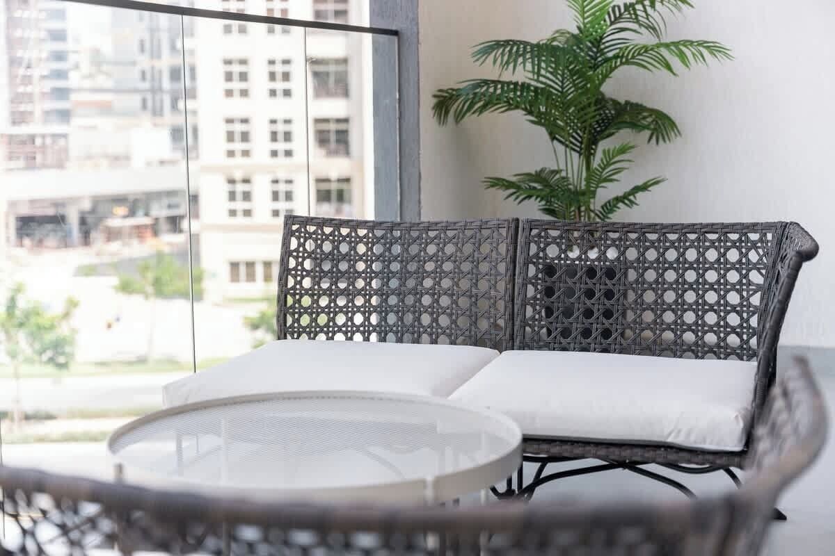 A balcony with a wicker sofa and a small round table, a potted plant, and a view of buildings in the background.