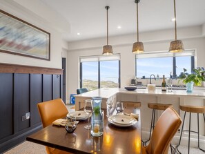 A modern dining area with a wooden table set for four, a kitchen in the background, and a view of the landscape through large windows.