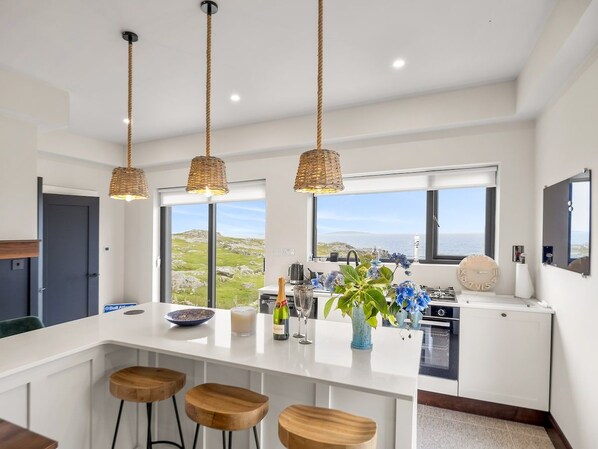 A modern kitchen with a large island, bar stools, hanging wicker pendant lights, and a view of the sea.
