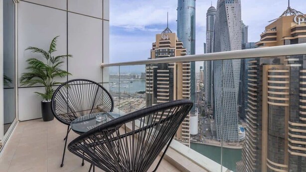 Un balcón con dos sillas de alambre negro y una mesita, con vistas a un paisaje urbano con edificios altos y una masa de agua.