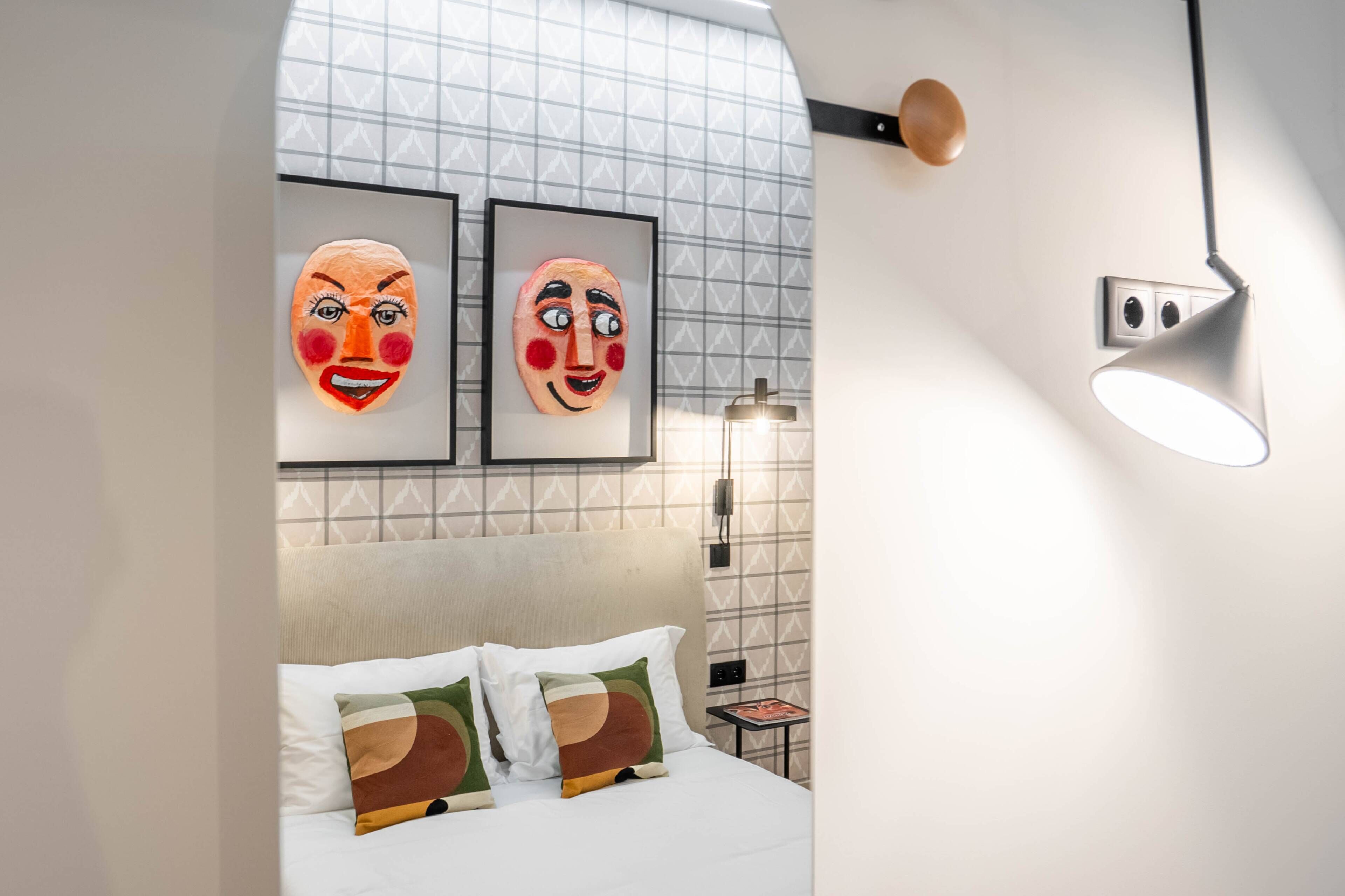 Una habitación de hotel moderna con una cama, dos almohadas, una mesita de noche, una lámpara y dos máscaras enmarcadas en la pared.