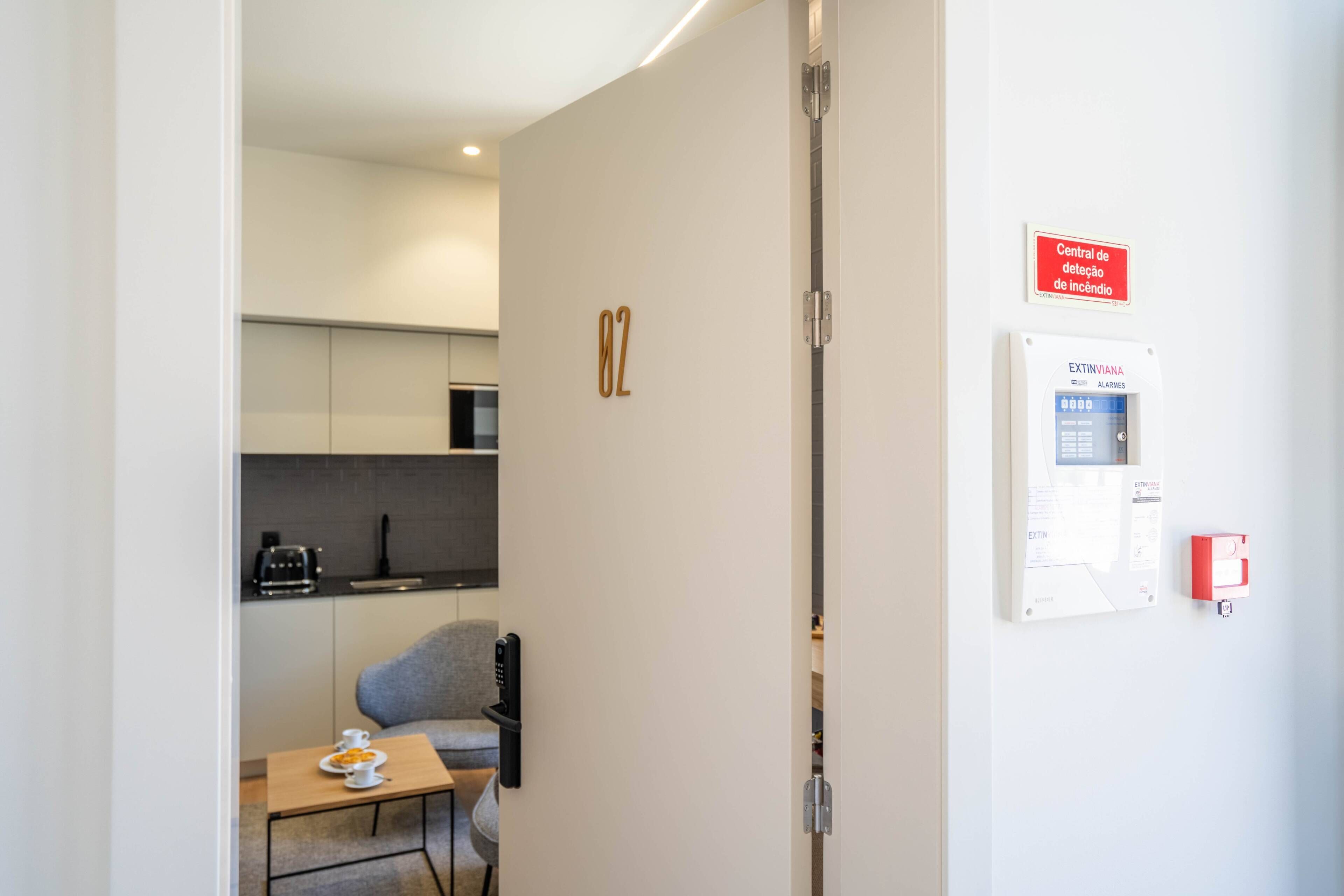 A modern kitchen with a toaster, a coffee maker, and a grey chair. A fire alarm and a control panel are mounted on the wall.