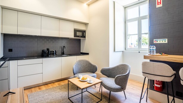 Eine moderne Küche mit weißen Schränken, einer Mikrowelle und einer Kaffeemaschine. Zwei graue Stühle stehen um einen kleinen Holztisch mit einer Speisenplatte und einer Tasse. Ein Fenster lässt natürliches Licht herein, und ein Feuerlöscher ist an der Wand sichtbar.