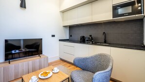 Eine moderne Küche mit weißen Schränken, einer Bosch-Mikrowelle und einer schwarzen Kaffeemaschine. An der Wand ist ein Flachbildfernseher angebracht, es gibt einen grauen Sessel und einen kleinen Holztisch mit einer Speiseplatte und einer Tasse.
