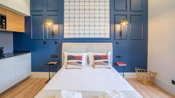 Ein modernes Hotelzimmer mit einem großen Bett, Blick durch ein Fenster, einer Küchenzeile und einem kleinen Tisch mit einem roten Buch.