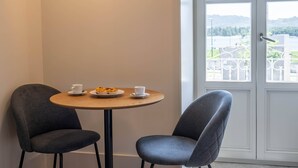 Une pièce d’angle avec une table ronde et deux chaises, une porte donnant sur une vue agréable, et une petite assiette avec de la nourriture et des tasses.