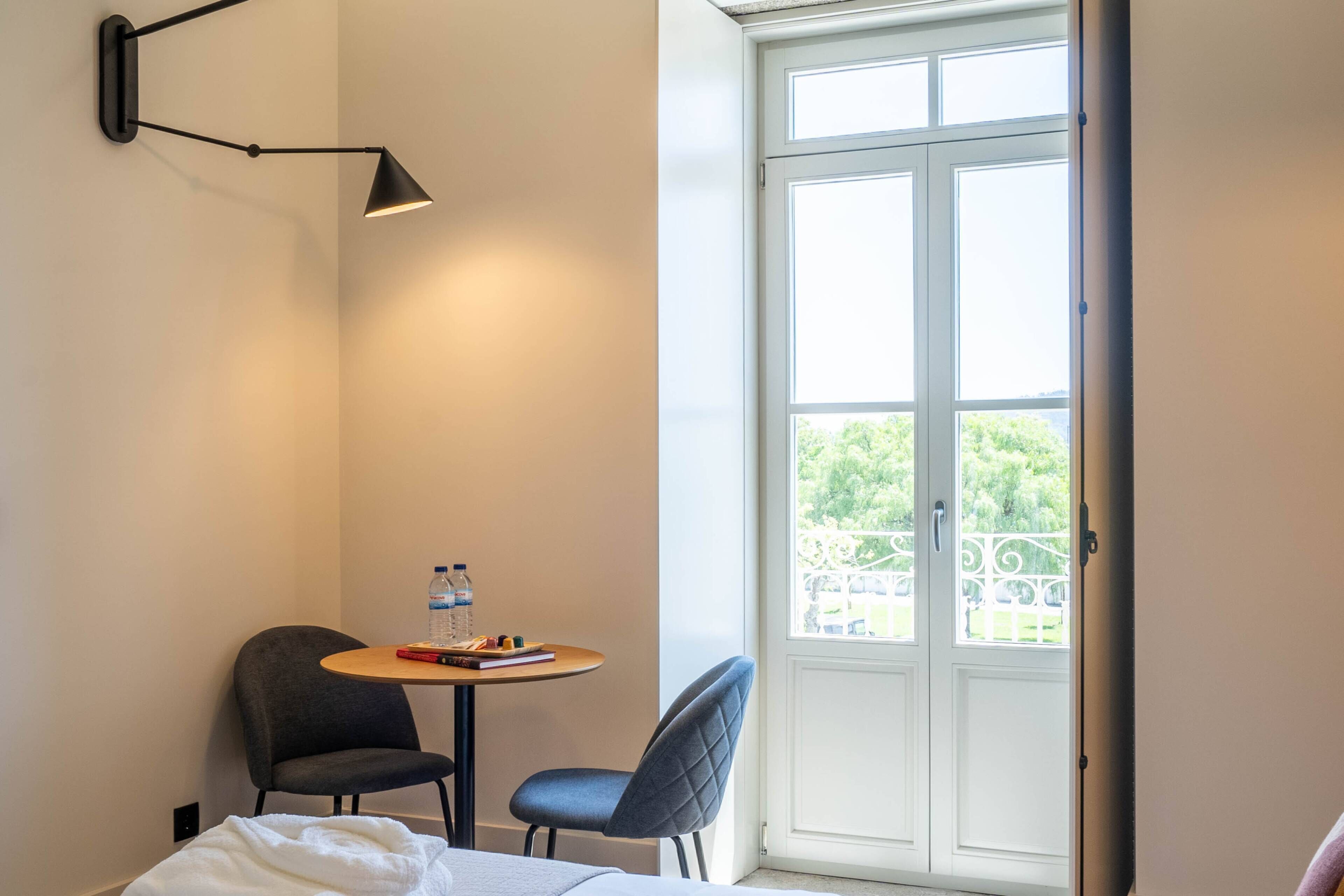 Ein Hotelzimmer mit einem Bett, einem kleinen Tisch mit Stuhl, einer Wasserflasche und einem Buch. Ein Fenster bietet Blick auf grüne Umgebung.