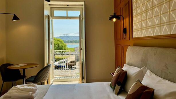 Une chambre d’hôtel comprenant un lit, un bureau, une chaise, un balcon avec vue sur un plan d’eau et des collines, ainsi qu’un mur décoré de motifs.