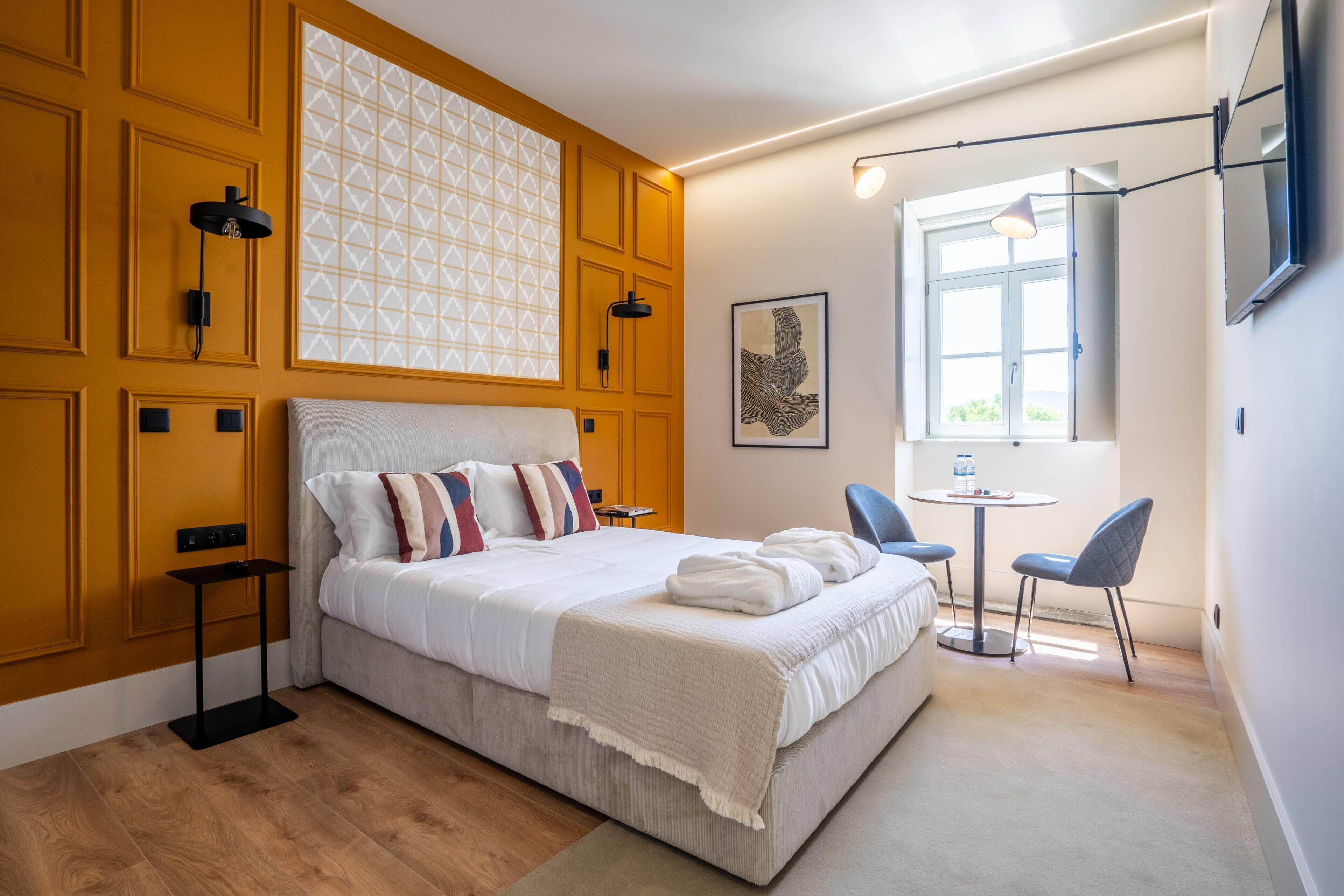 A modern bedroom with a large bed, bedside tables, a small table with chairs, a window, and a wall-mounted TV.