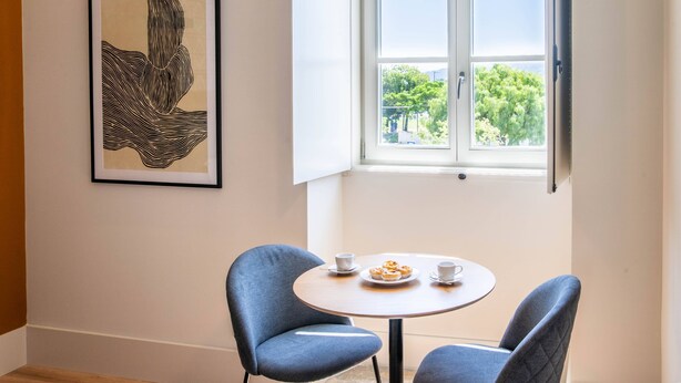 Una habitación con una mesa redonda, dos sillas, una obra de arte abstracta enmarcada y una ventana con vistas a árboles.
