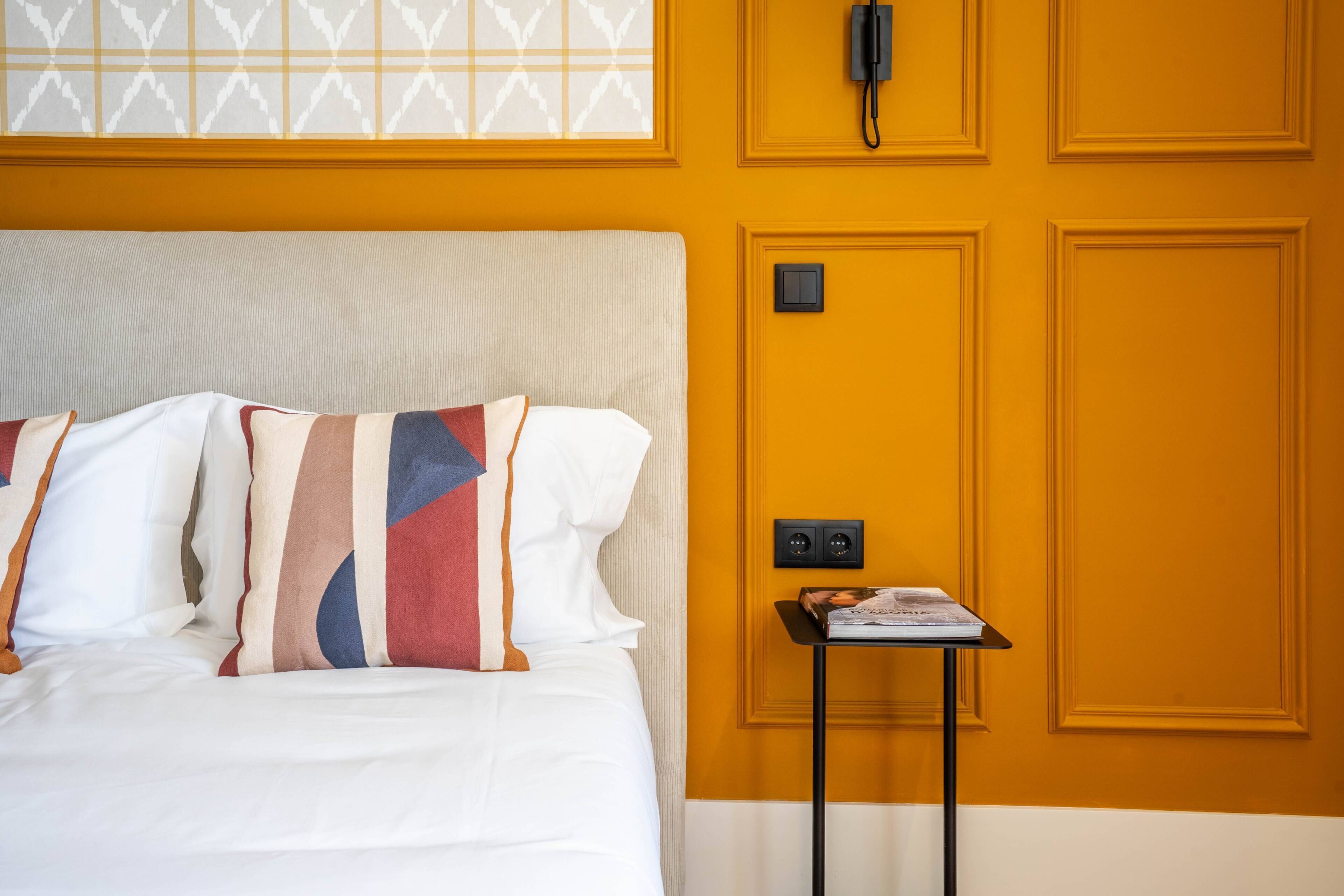 Ein Hotelzimmer mit einem Bett, einem Nachttisch mit einer Zeitschrift, einem Telefon und einem Lichtschalter an der Wand.