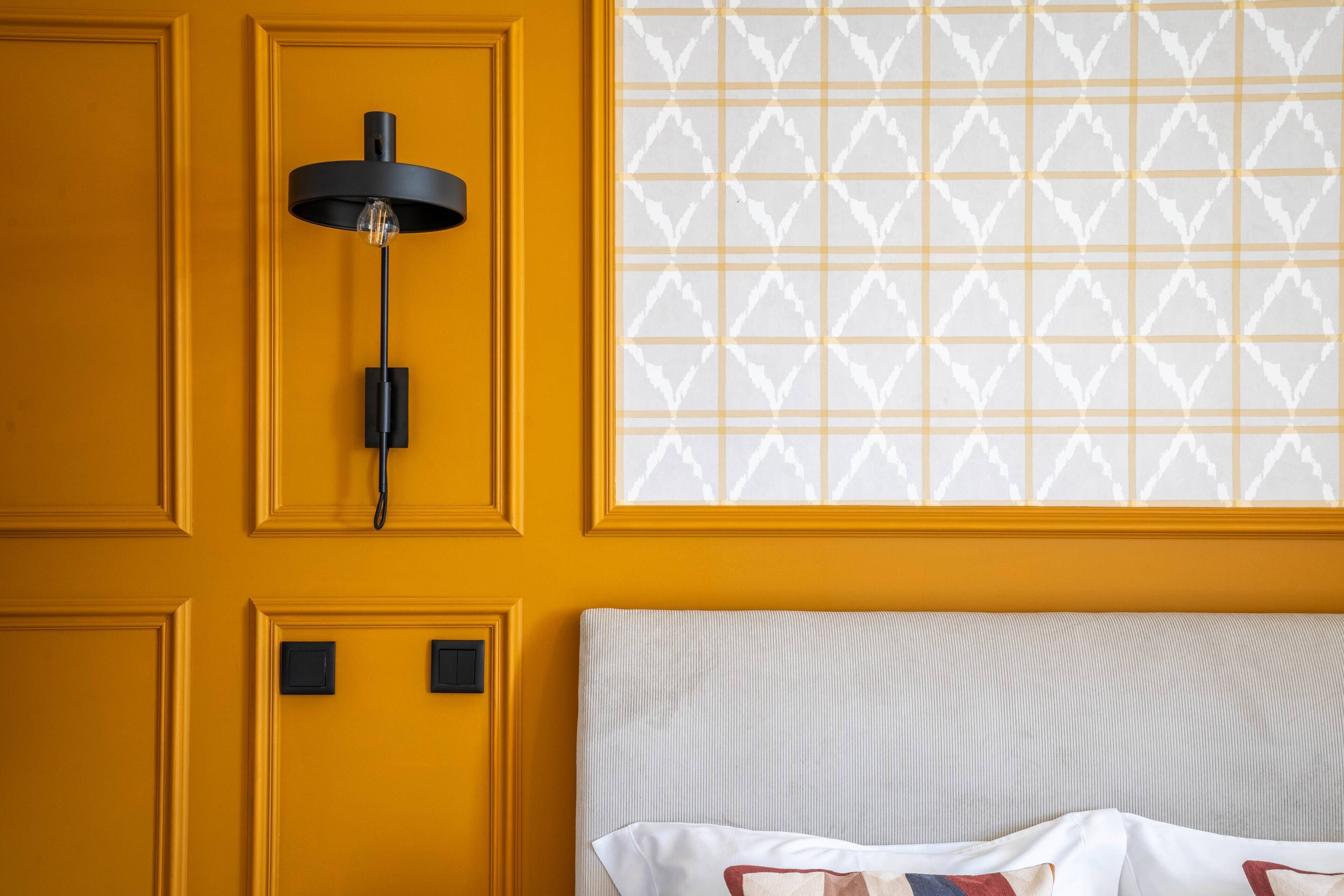 Una habitación con pared amarilla, lámpara de pared negra y ventana con diseño, además de cama con cabecera a rayas.