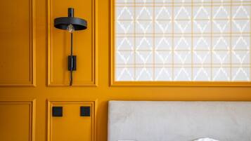 Una habitación con pared amarilla, lámpara de pared negra y ventana con diseño, además de cama con cabecera a rayas.