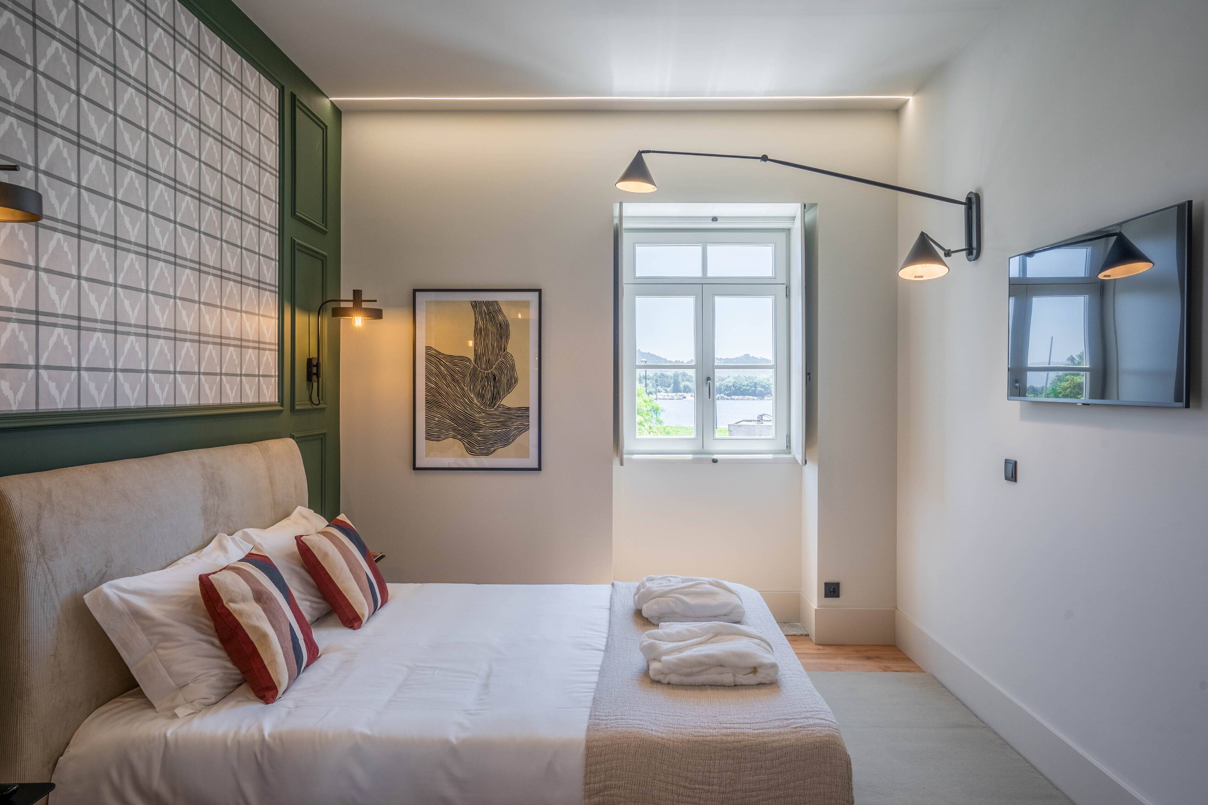 A bedroom with a bed, pillows, a window, a painting, and a wall-mounted lamp.