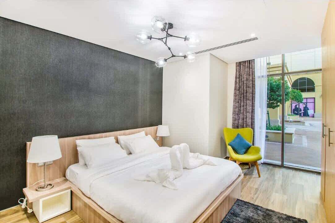 Una habitación de hotel moderna con una cama grande, una mesita de noche con lámpara, una silla de color amarillo brillante y vistas al exterior a través de un ventanal.
