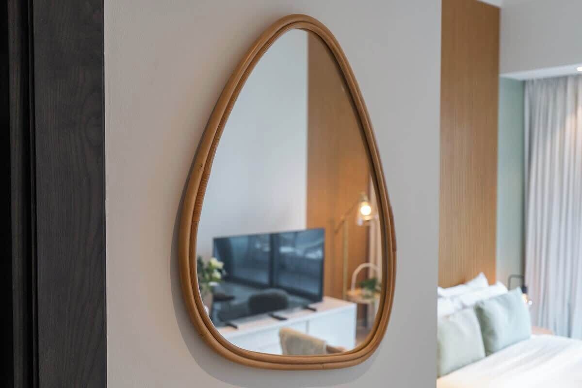 A hotel room with a large oval mirror on the wall, a bed with white bedding, a desk with a computer and a chair, and a television in the background.