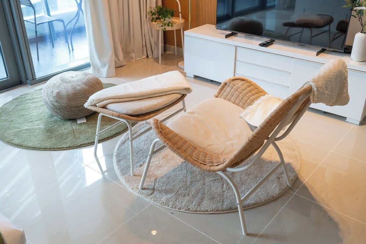 A modern living room with a wicker chair, a white sofa, a round rug, and a large window with a view of outdoor furniture.