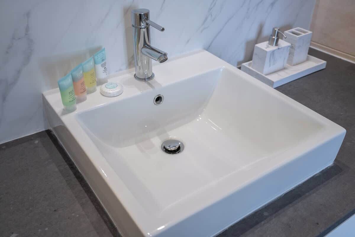 Ein weißes Waschbecken mit chromfarbenem Wasserhahn, eine Marmorwand und eine kleine Ablage mit Pflegeprodukten.