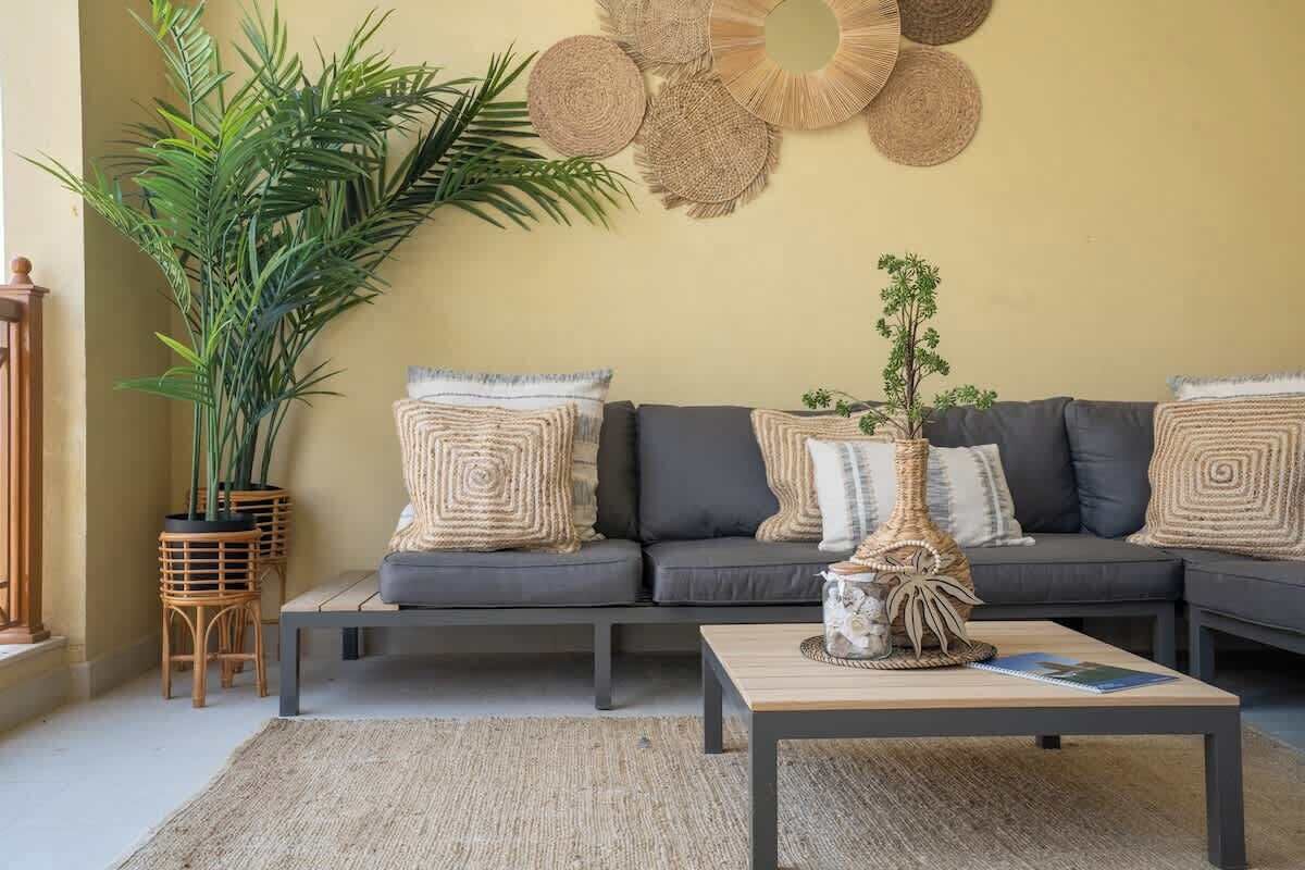 Una sala de estar con un sofá gris oscuro, cojines estampados, una mesa de centro trenzada y una planta grande en maceta.