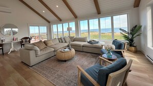 A spacious living room with a large sectional sofa, a round coffee table, and a view of the ocean through large windows. - Newly Built Ocean Front Cottage With Private Access To Small North Shore Beach (Park Corner)