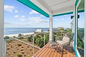Un balcón con vistas a la playa y el océano, una silla de mimbre y una terraza de madera.