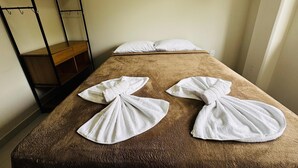 Un lit simple recouvert d’une couverture marron, deux serviettes blanches pliées posées dessus, et une étagère en métal sur le côté gauche.