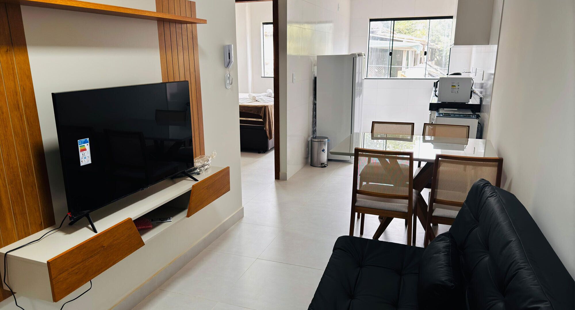 A modern living room with a flat-screen TV, wooden furniture, a dining area with a glass table, and a kitchen in the background.