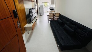 Un espace de vie compact comprenant un canapé noir, un téléviseur posé sur un meuble en bois, un coin cuisine et un espace repas.