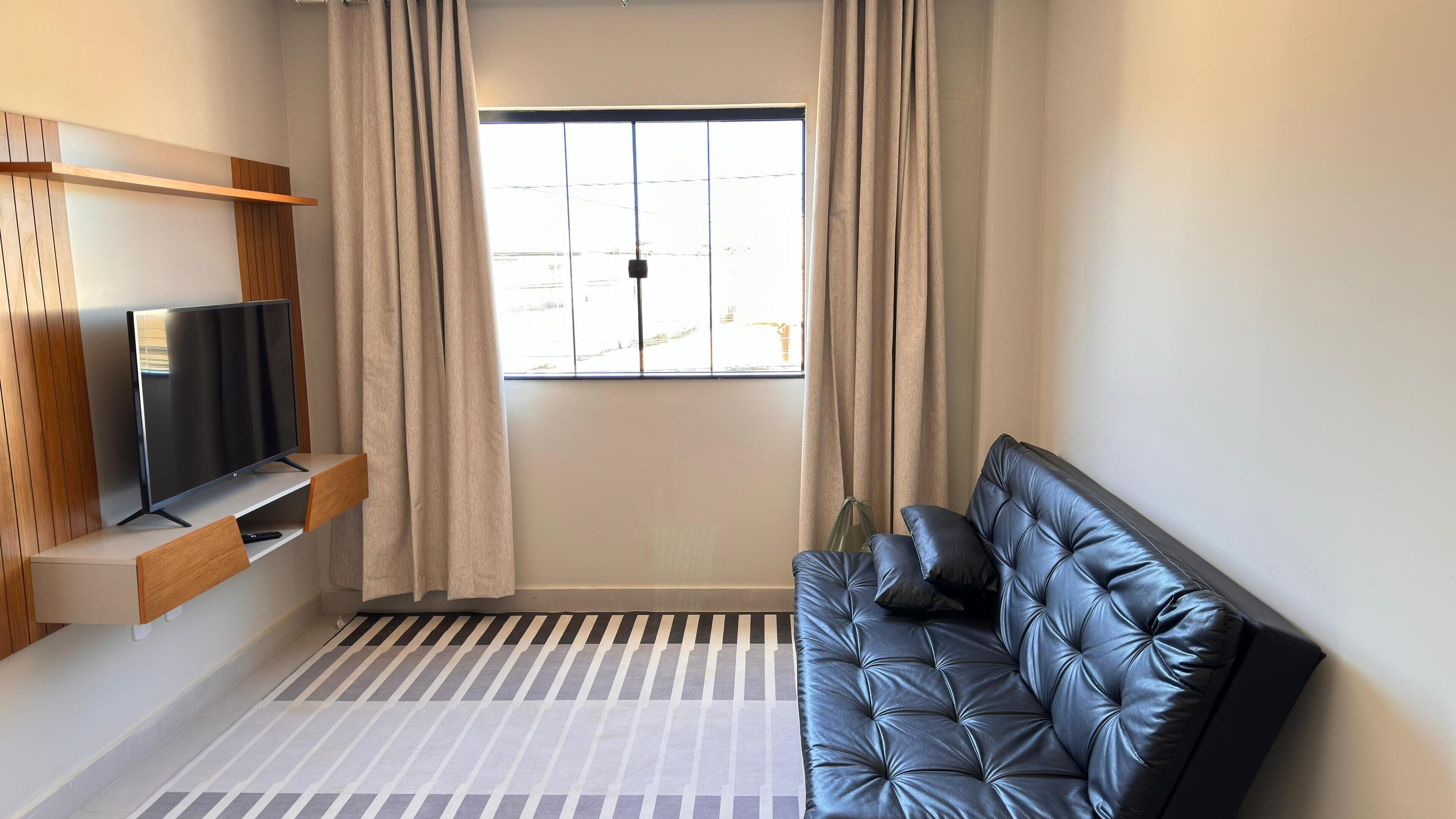 Ein modernes Wohnzimmer mit einer schwarzen Ledersofa, einem wandmontierten Fernseher und einem großen Fenster mit Vorhängen.