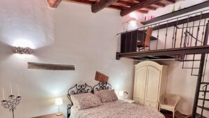Une chambre avec un parquet en bois disposé en chevrons, un lit avec une literie à motifs, une table de chevet avec une lampe, une commode et un balcon avec une balustrade.