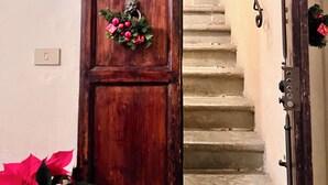 Une porte en bois ornée d’une guirlande décorative, un escalier avec une rampe en métal et un pot de fleurs contenant des fleurs rouges.